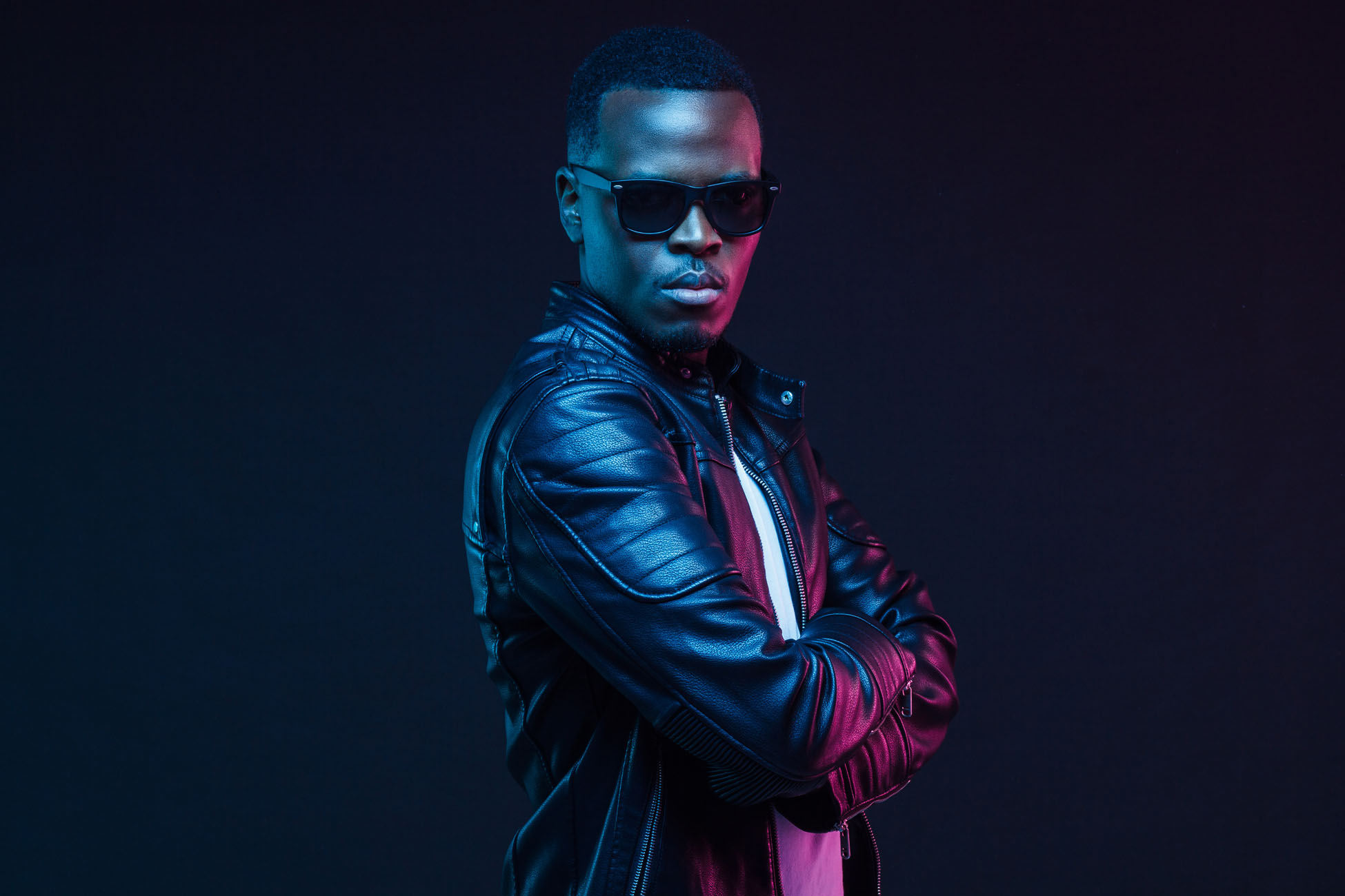 Studio neon portrait of stylish black man standing with crossed arms, wearing leather jacket and sunglasses